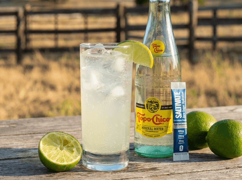 Glass of pale, lightly hazy ranch water with ice and a lime wedge beside a Topo Chico bottle and a Saltivate Raw Unflavored stick packet on a weathered wooden table