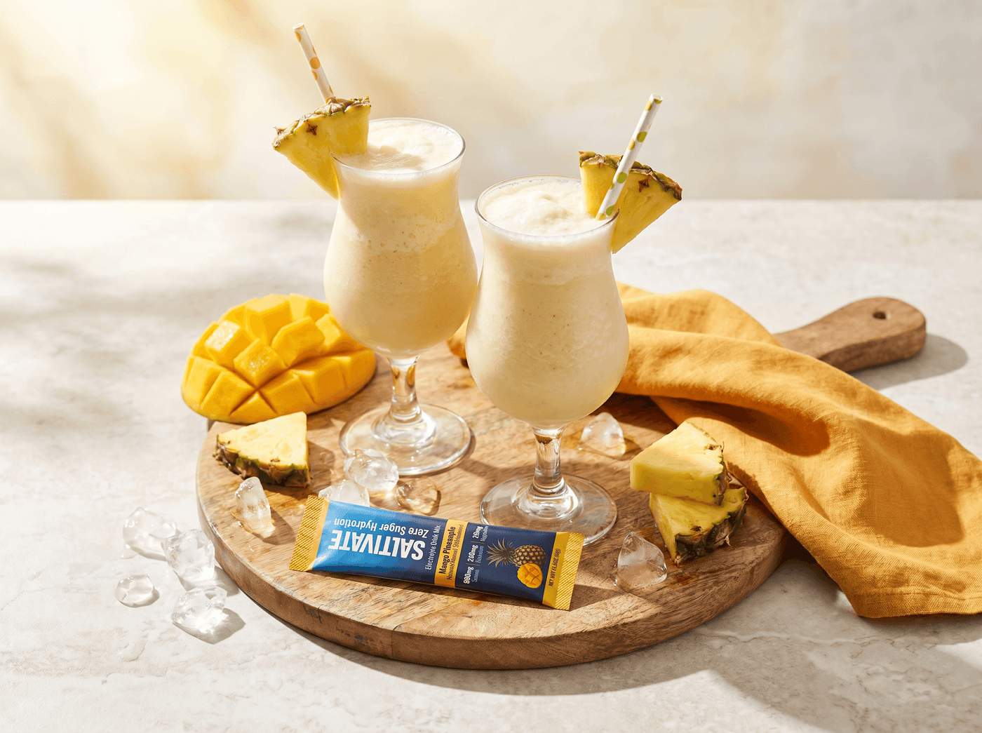 Two blended pina colada mocktails with pineapple wedges and a Saltivate Mango Pineapple stick packet