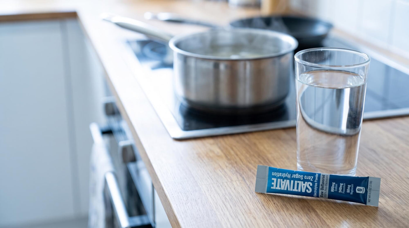 A sealed Saltivate Raw Unflavored electrolyte stick packet beside a glass of water on a kitchen counter with a pot of broth on the stove