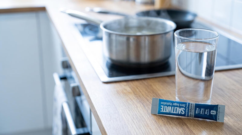 A sealed Saltivate Raw Unflavored electrolyte stick packet beside a glass of water on a kitchen counter with a pot of broth on the stove