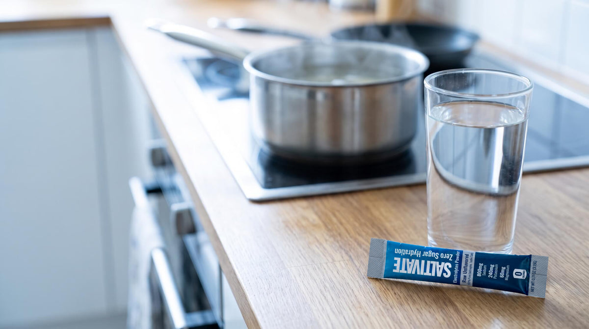 A sealed Saltivate Raw Unflavored electrolyte stick packet beside a glass of water on a kitchen counter with a pot of broth on the stove