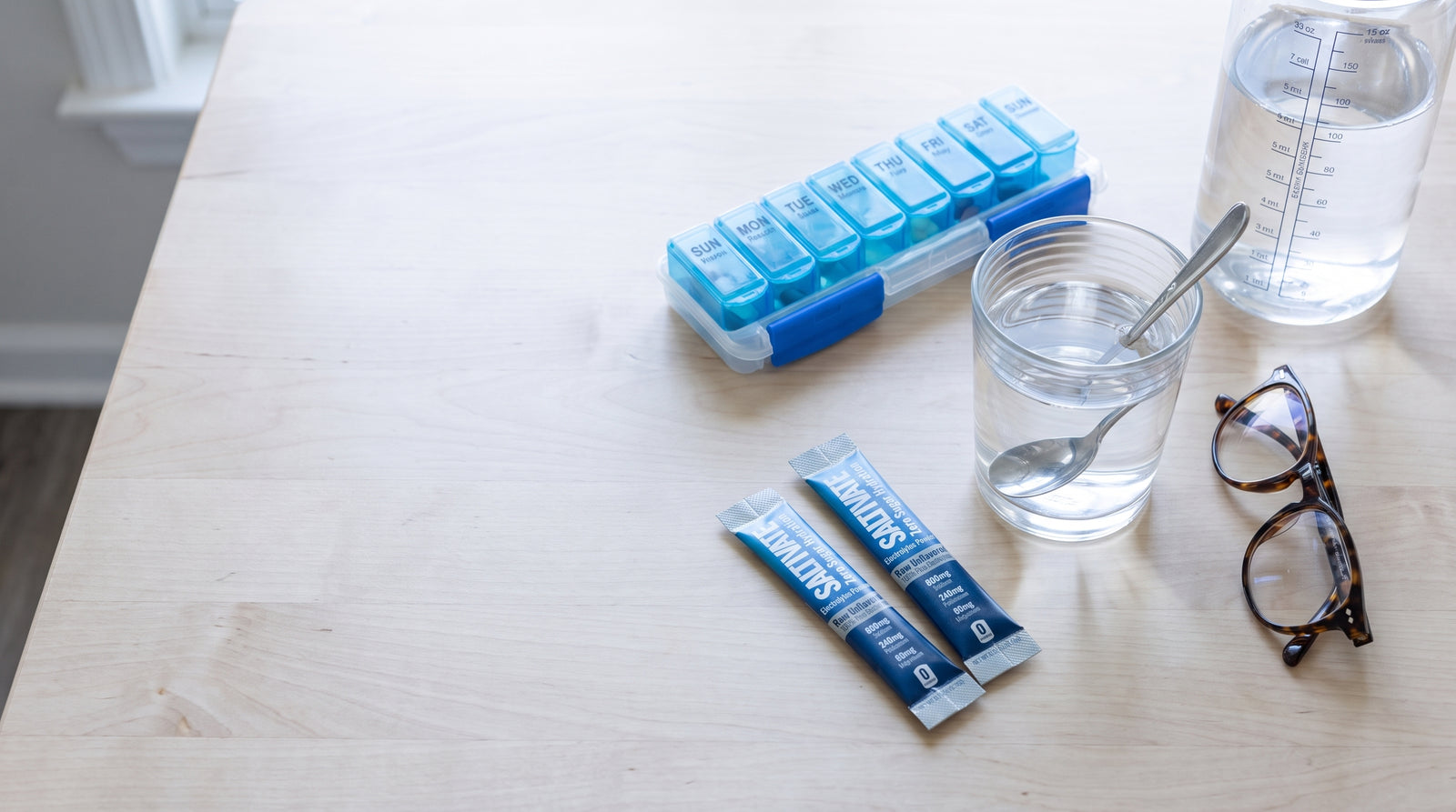 Kitchen table with a weekly pill organiser, a marked water bottle, a glass of water and two Saltivate Raw Unflavored stick packs