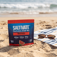 Saltivate Watermelon 30-count stick pack pouch standing in beach sand next to a striped towel and sunglasses