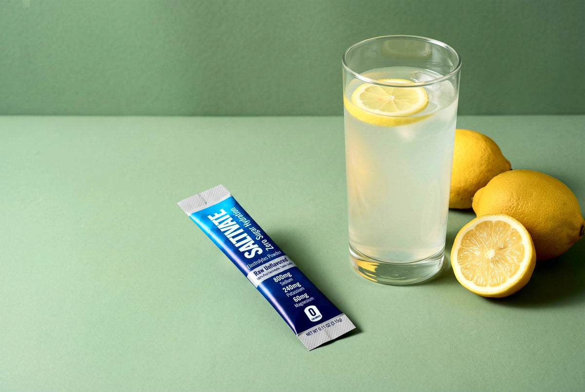 Saltivate Raw Unflavored stick pack beside a tall glass of hazy lemon water and lemons on a sage set