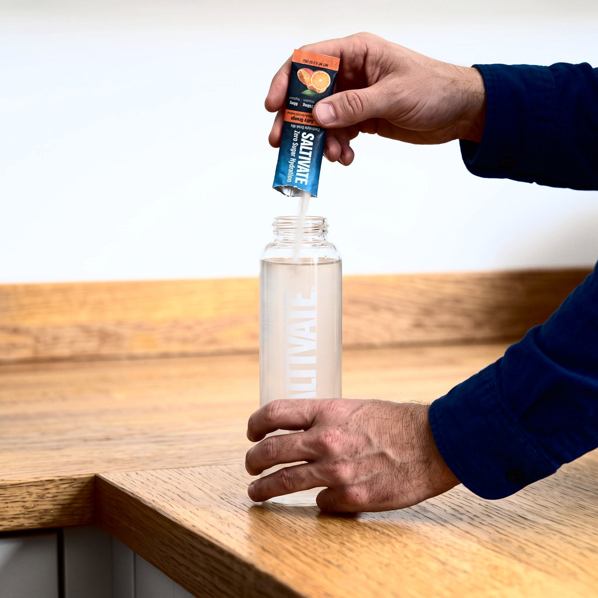 Hands pouring a Saltivate Salty Orange stick pack into a glass bottle of water on an oak counter