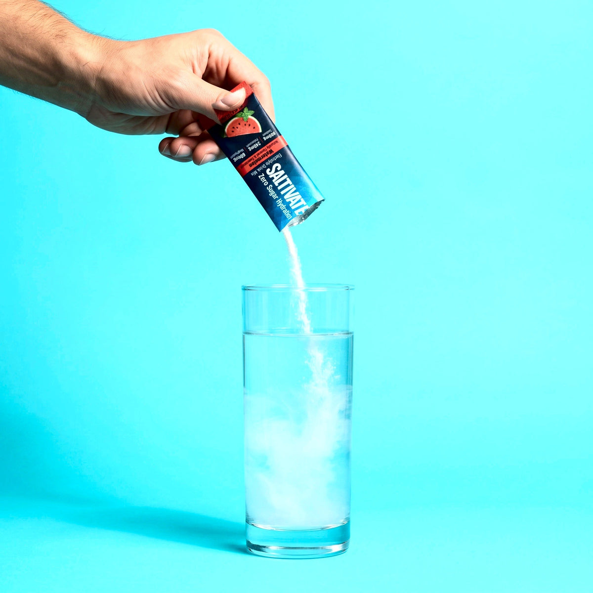 Hand pouring a Saltivate Watermelon stick pack into a tall glass of water, the powder clouding white, on an aqua set