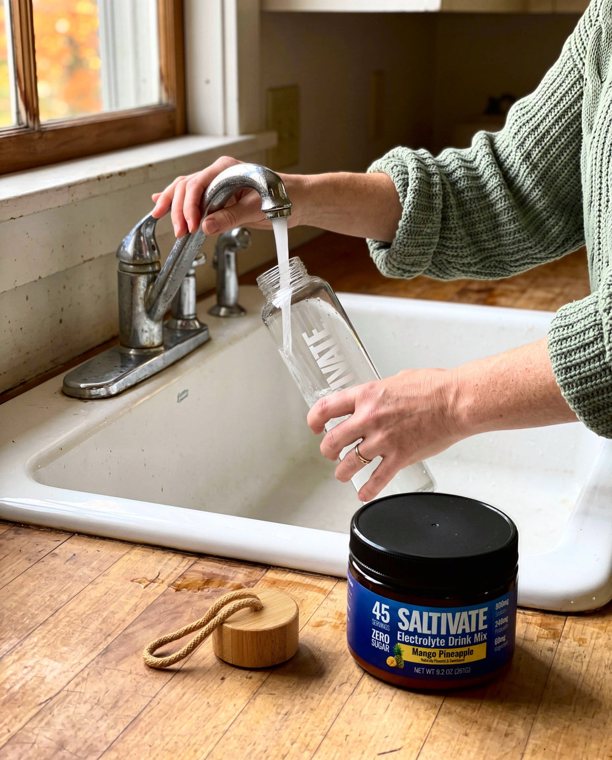 Hands filling a Saltivate glass bottle at a kitchen tap beside a Mango Pineapple jar