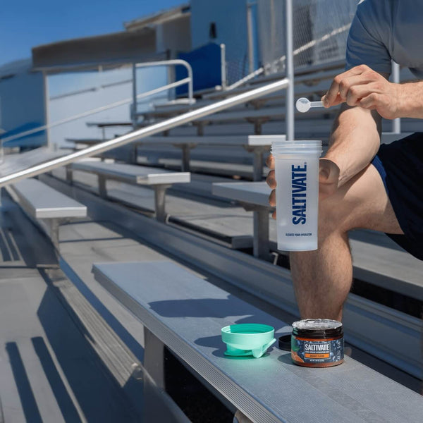 A person mixing Saltivate electrolyte powder into a shaker on stadium bleachers