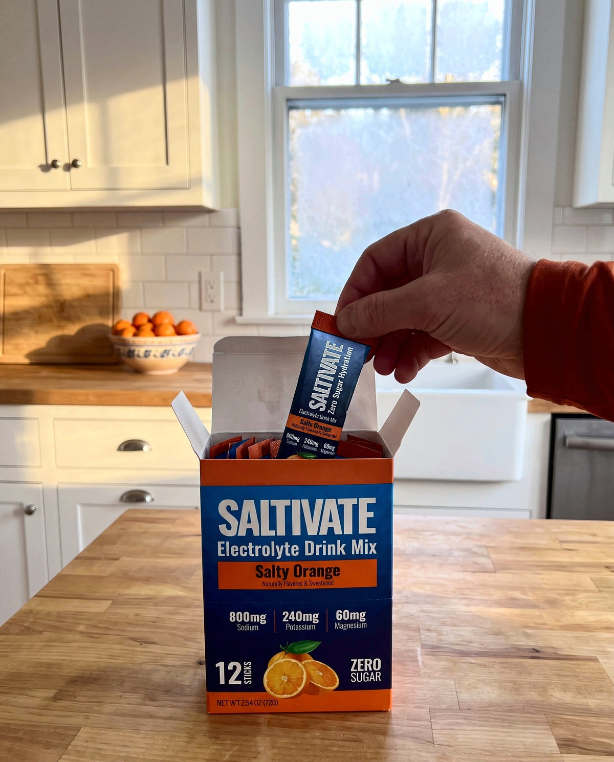 A hand lifting a Saltivate Salty Orange stick pack out of the open 12-stick carton on a butcher-block counter