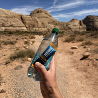 Hand holding a water bottle and a Saltivate Mango Pineapple stick on a desert hiking trail