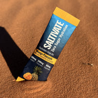 Saltivate Mango Pineapple electrolyte stick standing in rippled orange sand at sunset
