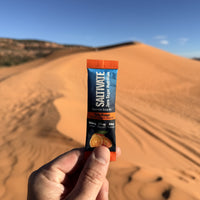Hand holding a Saltivate Salty Orange stick in front of orange sand dunes under a blue sky