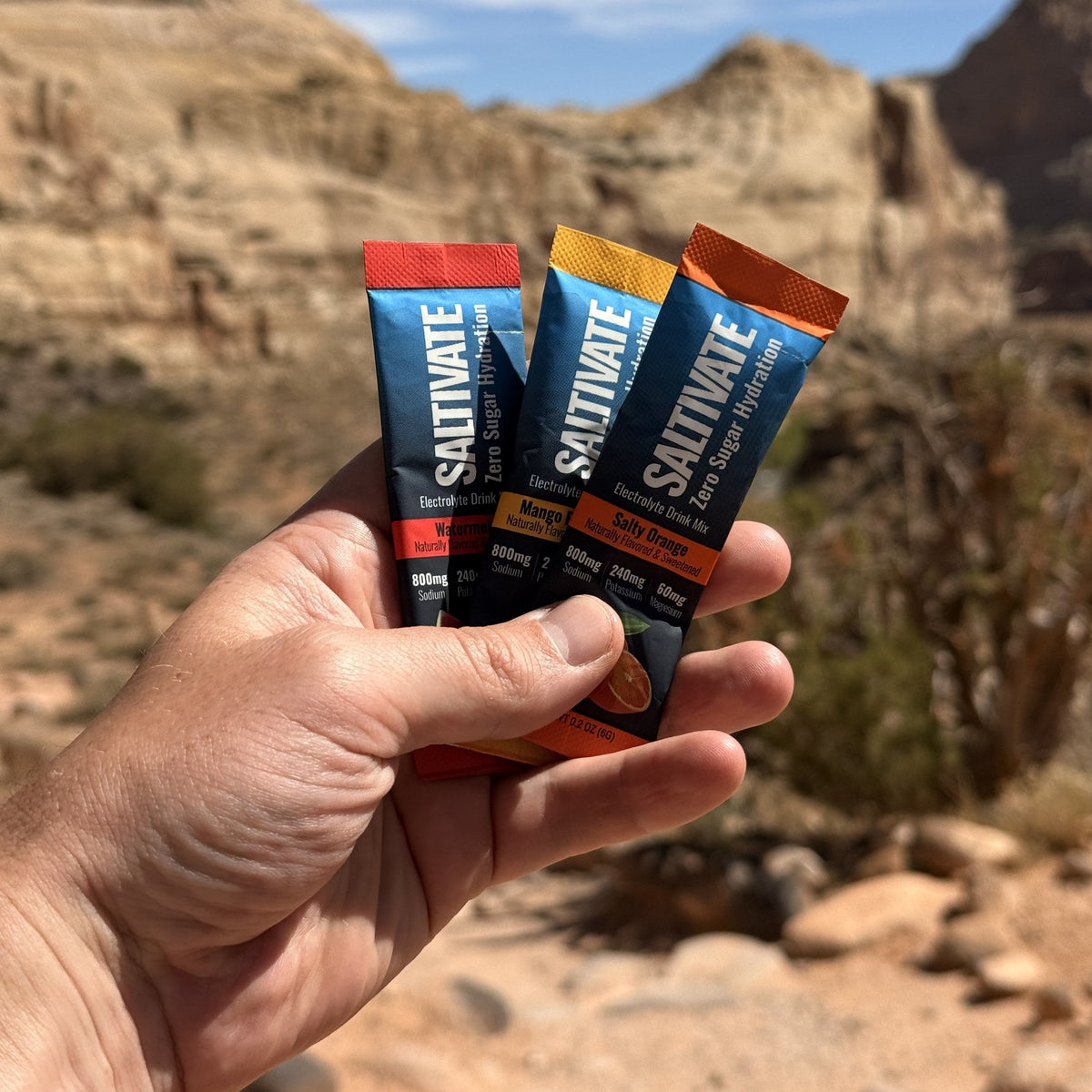 Hand holding Saltivate Watermelon, Mango Pineapple, and Salty Orange sticks in front of Utah sandstone cliffs