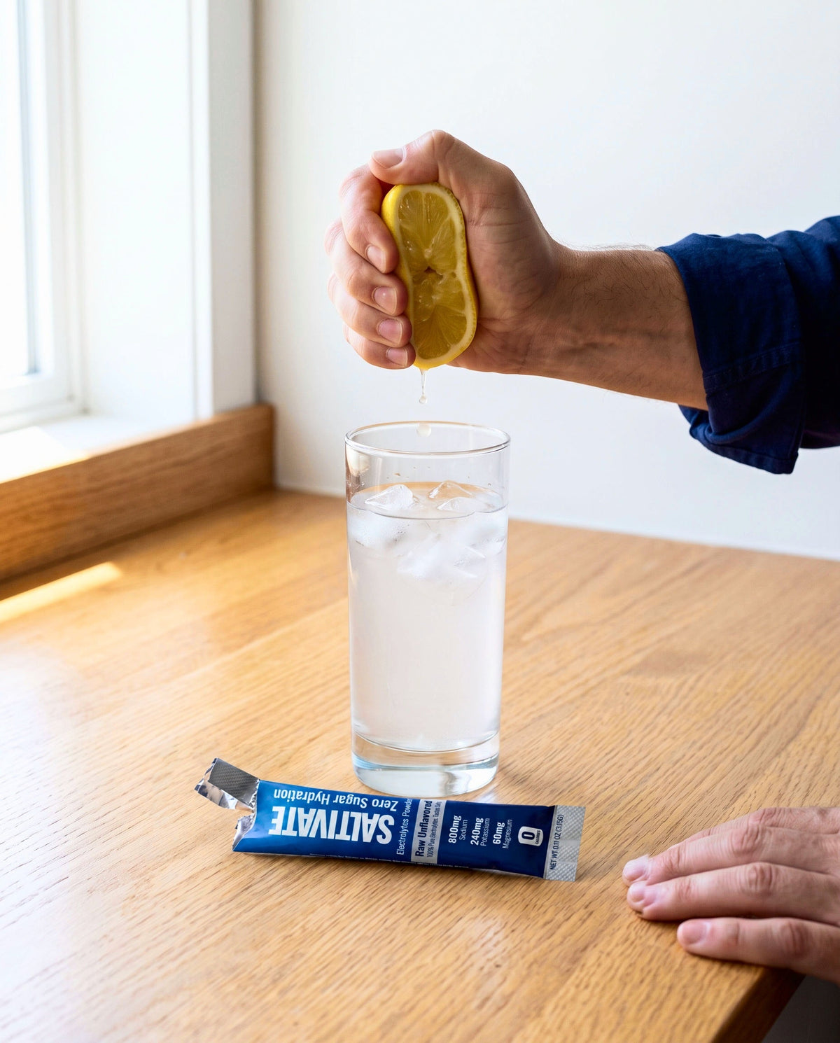 Hand squeezing a lemon into an iced glass beside a torn Saltivate Raw Unflavored stick pack