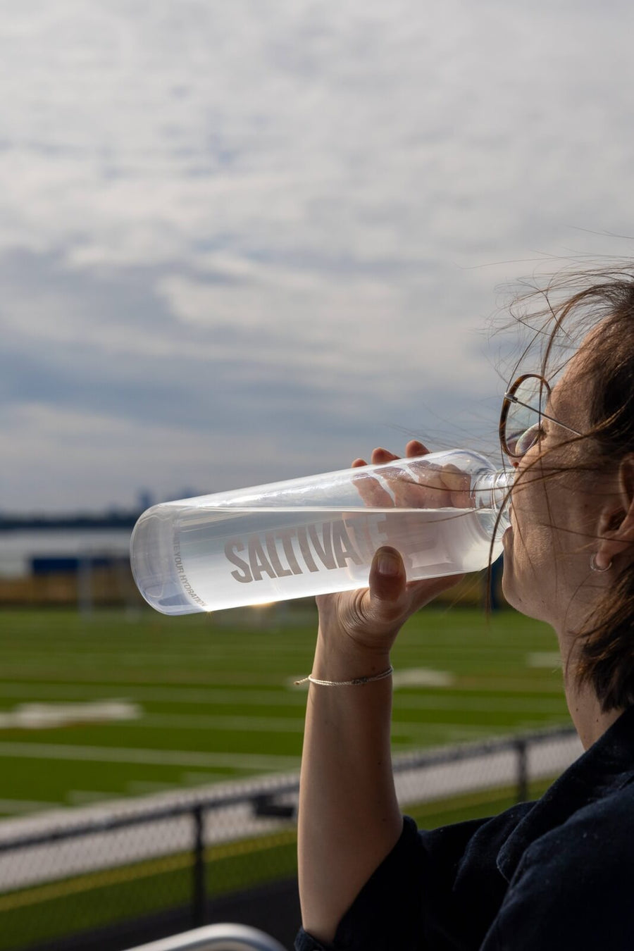A woman drinking from a Saltivate glass bottle, head tilted back under a cloudy sky