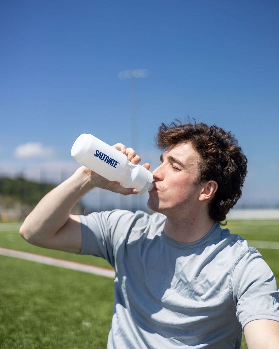 A young man drinking from a white Saltivate squeeze bottle on a sports field